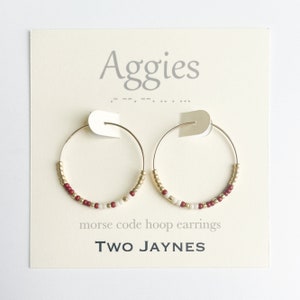 May include: Gold hoop earrings with red and white beads. The earrings are on a white card with the text "Aggies" and "morse code hoop earrings" and "TWO JAYNES".