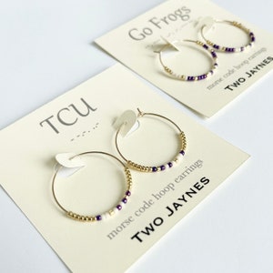 May include: Two pairs of gold hoop earrings with white and purple beads. The earrings are on cards with the text "TCU" and "Go Frogs" and the words "morse code hoop earrings" and "TWO JAYNES".