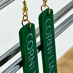 Puede incluir: Un par de pendientes de acr&iacute;lico verde con ganchos dorados. Los pendientes son rectangulares y tienen las palabras "Cornelia St" grabadas.