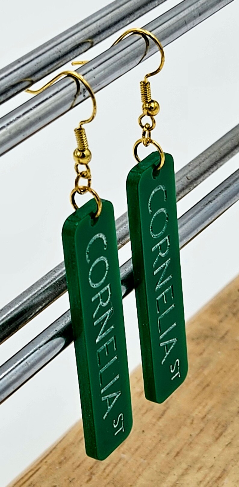 Puede incluir: Un par de pendientes de acr&iacute;lico verde con ganchos dorados. Los pendientes son rectangulares y tienen las palabras "Cornelia St" grabadas.