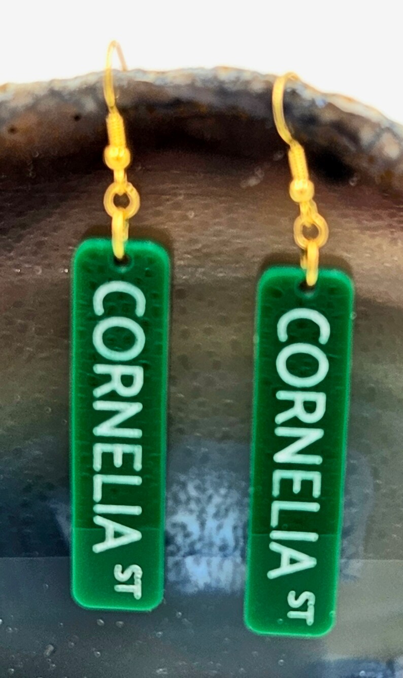 Puede incluir: Un par de pendientes de acr&iacute;lico verde con ganchos dorados. Los pendientes son rectangulares y tienen las palabras "Cornelia St" escritas en ellos.