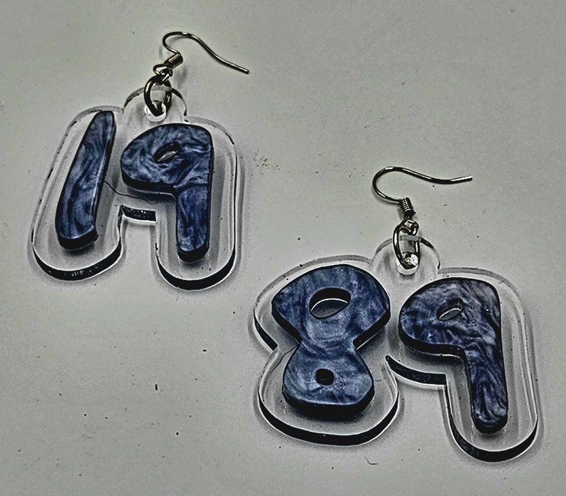 Pu&ograve; includere: Un paio di orecchini in acrilico trasparente con un design a marmi blu. Gli orecchini hanno la forma dei numeri 19 e 89.