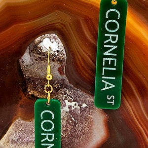 Puede incluir: Un par de pendientes de acr&iacute;lico verde con ganchos dorados. Los pendientes son rectangulares y tienen el texto "Cornelia St" impreso en blanco.