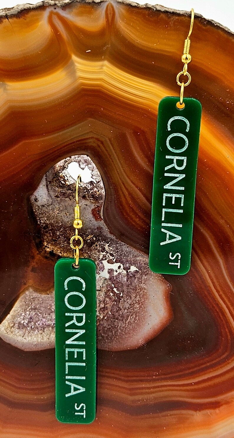 Puede incluir: Un par de pendientes de acr&iacute;lico verde con ganchos dorados. Los pendientes son rectangulares y tienen el texto "Cornelia St" impreso en blanco.