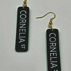Puede incluir: Un par de pendientes negros y verdes con ganchos dorados. Cada pendiente tiene un dije rectangular con el texto "Cornelia St" en letras blancas.