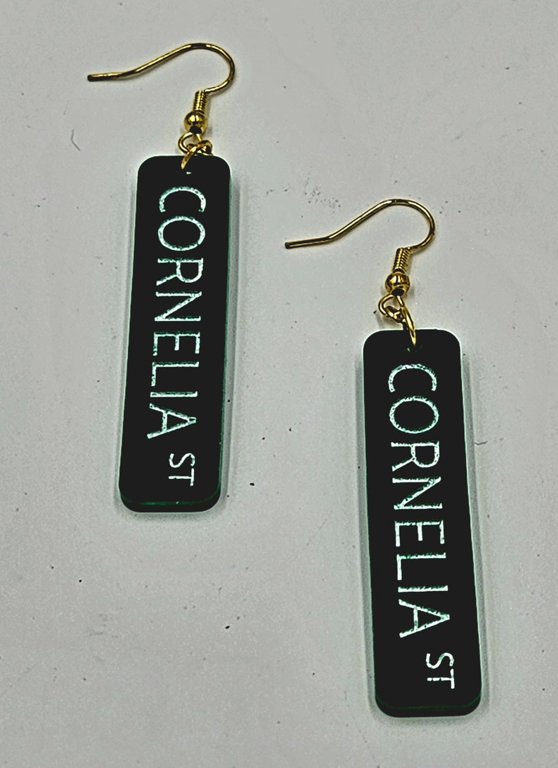 Puede incluir: Un par de pendientes negros y verdes con ganchos dorados. Cada pendiente tiene un dije rectangular con el texto "Cornelia St" en letras blancas.