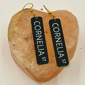 Puede incluir: Un par de pendientes de oro con dijes rectangulares negros que dicen "Cornelia St" en letras blancas. Los pendientes est&aacute;n sobre una roca rosa en forma de coraz&oacute;n.