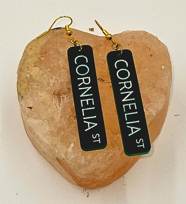 Puede incluir: Un par de pendientes de oro con dijes rectangulares negros que dicen "Cornelia St" en letras blancas. Los pendientes est&aacute;n sobre una roca rosa en forma de coraz&oacute;n.