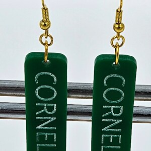 Puede incluir: Un par de pendientes de acr&iacute;lico verde con ganchos dorados. Los pendientes son rectangulares y tienen las palabras "Cornelia St" escritas en blanco.