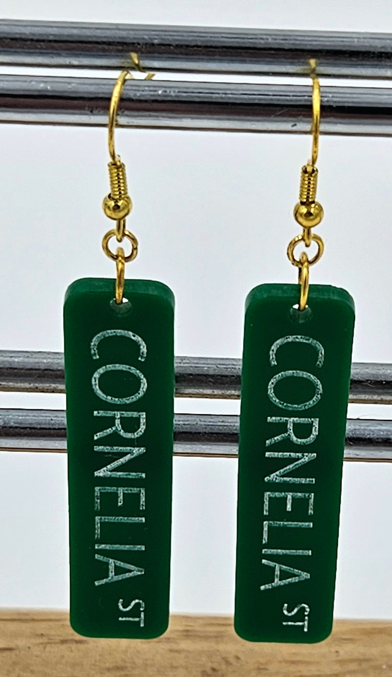 Puede incluir: Un par de pendientes de acr&iacute;lico verde con ganchos dorados. Los pendientes son rectangulares y tienen las palabras "Cornelia St" escritas en blanco.