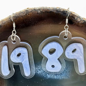 Pu&ograve; includere: Un paio di orecchini in acrilico trasparente con ganci argentati. Gli orecchini hanno la forma dei numeri 19 e 89. I numeri sono riempiti con un design marmoreo blu chiaro.