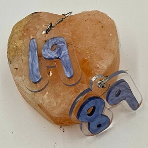 Pu&ograve; includere: Un paio di orecchini in acrilico trasparente con un design a marmi blu. Gli orecchini hanno la forma dei numeri 1 e 9. Gli orecchini sono appesi a una roccia rosa a forma di cuore.
