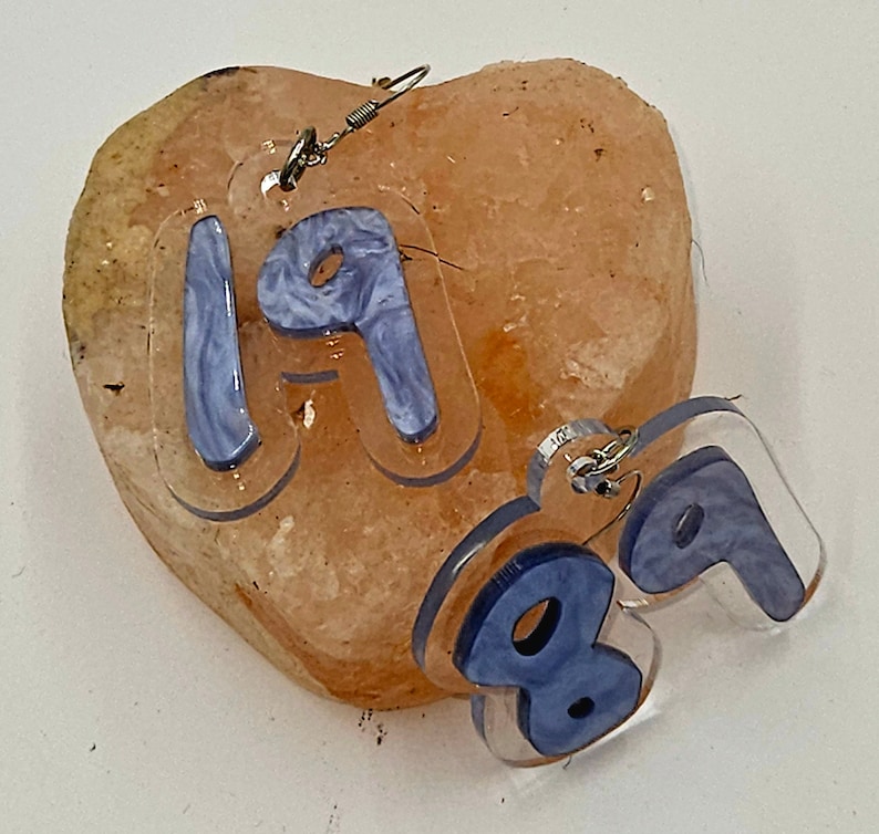 Pu&ograve; includere: Un paio di orecchini in acrilico trasparente con un design a marmi blu. Gli orecchini hanno la forma dei numeri 1 e 9. Gli orecchini sono appesi a una roccia rosa a forma di cuore.