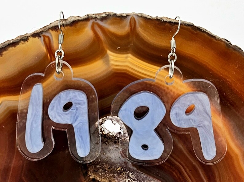 Pu&ograve; includere: Un paio di orecchini in acrilico trasparente con ganci argentati. Gli orecchini hanno la forma dei numeri 1989, con un design a spirale blu chiaro all'interno di ogni numero.