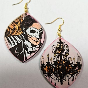 Puede incluir: Un par de pendientes colgantes con fondo rosa. Un pendiente presenta una ilustración en blanco y negro de un hombre con máscara. El otro pendiente presenta una ilustración en negro y dorado de una lámpara de araña.