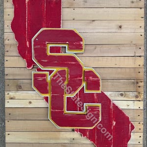 May include: A wooden sign featuring the outline of California in red, with the letters "SC" in a layered design. The letters are red with yellow and white accents. The sign is mounted on a wooden plank background.