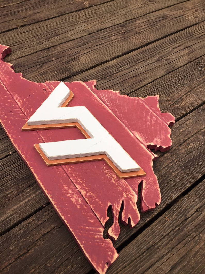 Puede incluir: Un letrero de madera rojo con forma del estado de Virginia con una "F" blanca en el centro. El letrero est&aacute; desgastado y tiene un aspecto r&uacute;stico.