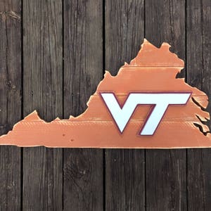 Puede incluir: Un recorte de madera del estado de Virginia pintado en color naranja quemado con un logotipo "VT" blanco en el centro. El logotipo tiene un contorno marr&oacute;n.