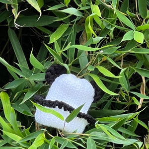 May include: A handmade white and black crochet panda hat with black ears and a black band. The hat is nestled among green bamboo leaves, creating a playful and whimsical scene. The hat is a unique accessory.