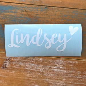 May include: A light blue decal with the name "Lindsey" in white cursive script, accompanied by a white heart. The decal is placed on a wooden surface with a distressed blue and brown finish.
