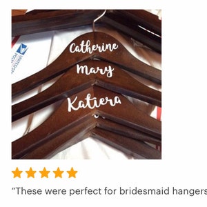 May include: Dark brown wooden hangers with white cursive lettering. The names "Catherine", "Mary", and "Katiera" are displayed on the hangers. The hangers are designed for clothing and are likely for a wedding or special event.