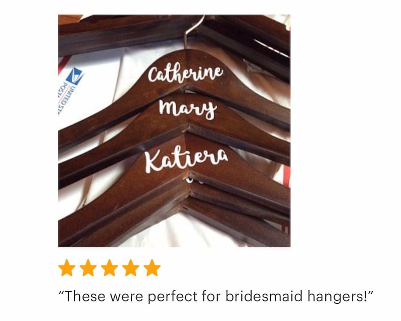 May include: Dark brown wooden hangers with white cursive lettering. The names "Catherine", "Mary", and "Katiera" are displayed on the hangers. The hangers are designed for clothing and are likely for a wedding or special event.