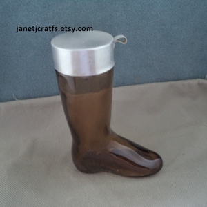 May include: A brown glass boot-shaped bottle with a silver lid. The bottle is empty and has a vintage look.