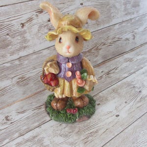 May include: A figurine of a bunny wearing a yellow hat and purple vest with orange buttons. The bunny holds a basket of red items and a bouquet of pink flowers. It stands on a green base with red roses.