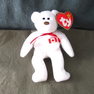 May include: White plush teddy bear with a red ribbon and a Canadian flag patch. The bear has a red nose and black eyes. The bear is wearing a red "ty" heart tag with a gold star.