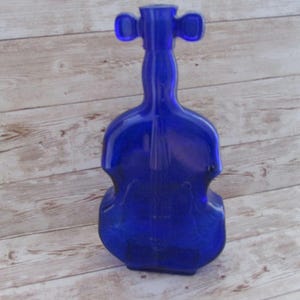 May include: A cobalt blue glass bottle shaped like a violin. The bottle has a long neck with two rounded protrusions at the top. The body of the bottle has the curved shape of a violin. The bottle is sitting on a light wood surface.