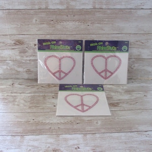 May include: Three packages of pink iron-on rhinestone studs in the shape of a peace sign inside a heart. The packages are labeled "Iron-On Rhinestone Studs".
