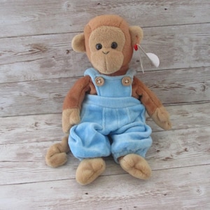 May include: A plush monkey toy with brown fur and a light blue overall. The monkey has a tan face, ears, hands, and feet. The overalls have wooden button closures. A white tag is attached to the monkey's ear.