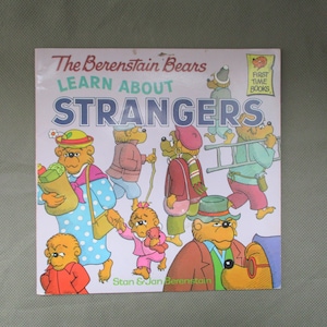 May include: The Berenstain Bears Learn About Strangers book cover features a group of bears in various situations, including a bear carrying groceries, a bear climbing a ladder, and a bear holding a teddy bear. The book is part of the First Time Books series.