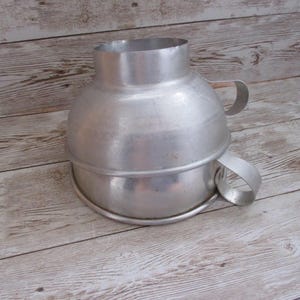 May include: A vintage, aluminum funnel with a wide mouth and a handle on each side. The funnel has a cylindrical top and a flared, rounded body. The object is likely used for pouring liquids or dry ingredients.