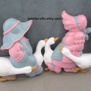 May include: Two vintage wall decor figurines of children riding geese. The girl is wearing a pink dress and blue bonnet, and the boy is wearing blue overalls and a blue bonnet. Both figurines are painted in pastel colors.