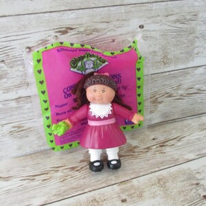 May include: A Cabbage Patch Kids doll in a pink dress with a white collar, black shoes, and brown hair. The doll is in its original packaging, which is pink and green and has the words "Cabbage Patch Kids" on it.
