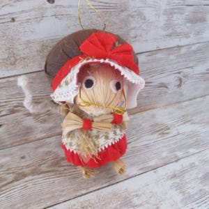 May include: A handmade straw doll ornament with a brown hat, red bow, and red dress. The doll has a straw face with black eyes and gold wire glasses. The ornament is suspended by a gold string, perfect for holiday decor.