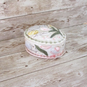 May include: Oval ceramic trinket box with a lid, featuring a seashell and botanical design. The box is primarily white with pink, green, and yellow accents. The lid has a raised design of leaves and shells. The box is on a light wood surface.