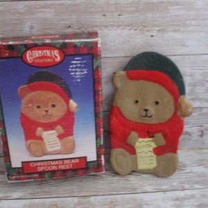 May include: A Christmas bear spoon rest and its box. The bear is wearing a red sweater and a green and red hat. The bear is holding a scroll. The box has the words "Christmas Collectibles" and "Christmas Bear Spoon Rest".