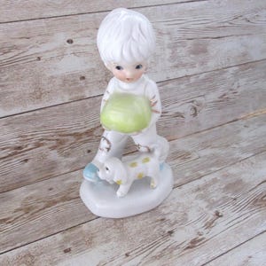May include: A ceramic figurine of a child holding a green ball, with a small dog at their feet. The child and dog are white with blue accents. The figurine is on a white base.