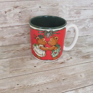 May include: A red ceramic mug with a white handle and rim. The mug features a festive design of two brown teddy bears holding hands under mistletoe. The interior of the mug is dark green.