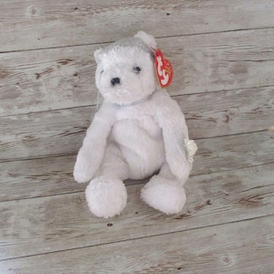 May include: A white plush teddy bear with soft fur, black plastic eyes, and a black stitched nose. The bear features a red heart-shaped tag attached to its ear with the white text ty printed on it, and a small white fabric flower on its left arm.