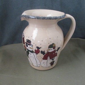 May include: A ceramic pitcher with a cream-colored glaze and a gray rim. The pitcher features a winter scene with two snowmen, a bird, and snowflakes.