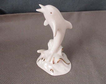 May include: A white porcelain figurine of a dolphin leaping out of the water. The dolphin is painted with gold accents and is set on a base with a wave design.