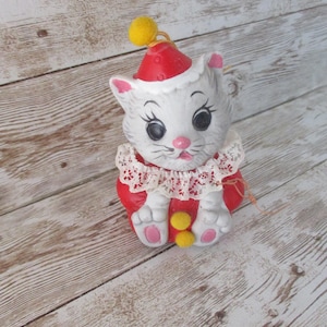 May include: A ceramic cat figurine in a clown costume. The cat is white with a red hat and outfit, a lace collar, and yellow pom-poms. The cat has large black eyes, pink ears, and a pink nose. The figurine is sitting on a wooden surface.