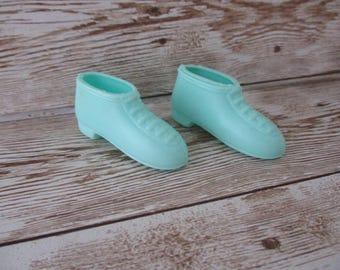 May include: A pair of light blue doll shoes. The shoes are a solid color with a rounded toe and a ridged design on the top. The shoes are sitting on a wooden surface.