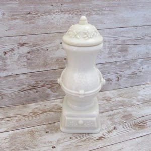 May include: A white ceramic jar with a lid, featuring a floral design on the lid and a decorative base. The jar has a unique shape with a bulbous midsection and a small knob on top. The overall design is elegant and classic.