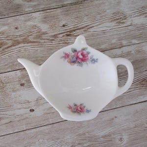 May include: White ceramic teapot-shaped tea bag holder with a floral design. The holder features a spout, handle, and a small knob on top. The floral design includes pink and blue flowers. The holder is placed on a light brown wooden surface.