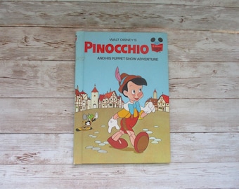 May include: Vintage Walt Disney's Pinocchio book. The cover features Pinocchio in a red outfit with a yellow shirt and blue bow tie, walking in front of a town. The title is in red and the background is light blue.