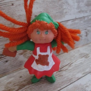May include: A vintage doll with orange yarn hair, green hat and shoes, and a red dress with a white apron featuring a teddy bear. The doll is holding a small brown object.